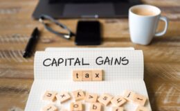 Closeup on notebook over vintage desk surface, front focus on wooden blocks with letters making Capital Gains Tax text. Business concept image with office tools and coffee cup in background