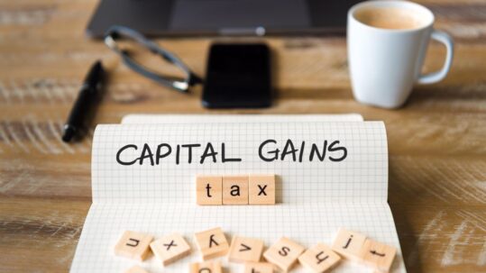 Closeup on notebook over vintage desk surface, front focus on wooden blocks with letters making Capital Gains Tax text. Business concept image with office tools and coffee cup in background